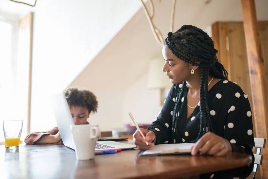 A mother working with a notebook and laptop while her child sits beside her, with a small red accent on a pen